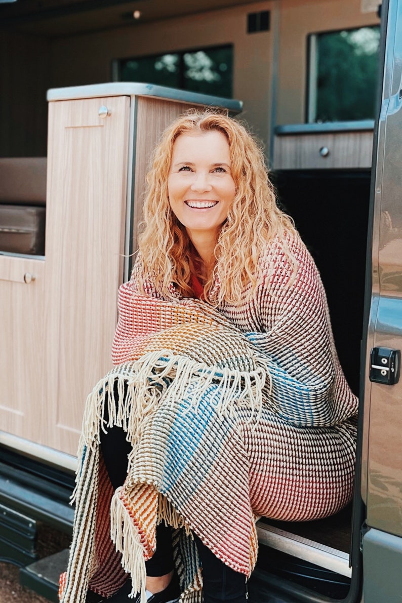 Pu&ograve; includere: Una donna seduta all'interno di un camper, avvolta in una coperta a righe colorate con frange. Sta sorridendo e guardando la telecamera.