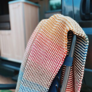 Pu&ograve; includere: Una coperta di maglia a righe multicolore drappeggiata su una sedia pieghevole nera e blu. La coperta ha strisce di arancione, rosso, marrone, blu e bianco.