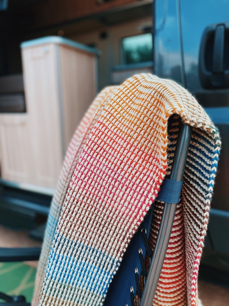 Pu&ograve; includere: Una coperta di maglia a righe multicolore drappeggiata su una sedia pieghevole nera e blu. La coperta ha strisce di arancione, rosso, marrone, blu e bianco.