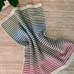 May include: A striped crochet blanket with a gradient of colours, starting with green and transitioning to blue, then brown, and finally ending with red. The blanket is folded and lying on a woven rug.