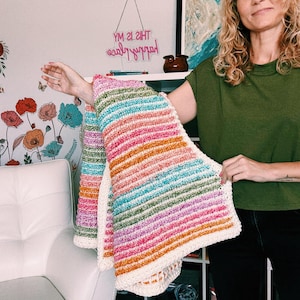 Op de afbeelding: Een handgemaakte, kleurrijke gehaakte deken met horizontale strepen in tinten roze, oranje, groen en blauw. De deken heeft een cr&egrave;mekleurige rand. De tekst "YM 2I 2IHT" is zichtbaar op de achtergrond.