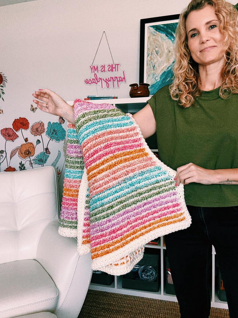 Op de afbeelding: Een handgemaakte, kleurrijke gehaakte deken met horizontale strepen in tinten roze, oranje, groen en blauw. De deken heeft een cr&egrave;mekleurige rand. De tekst "YM 2I 2IHT" is zichtbaar op de achtergrond.