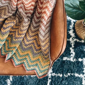 Pu&ograve; includere: Una coperta all'uncinetto con un motivo a chevron in tonalit&agrave; di marrone, arancione, giallo, verde e viola. La coperta &egrave; drappeggiata su una poltrona in pelle marrone.