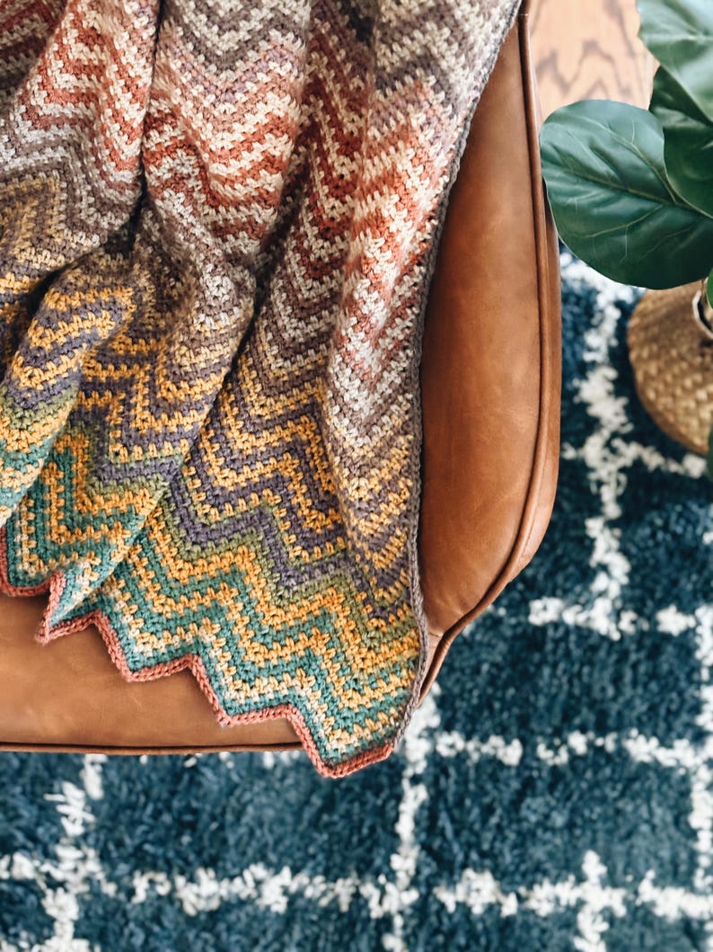 Pu&ograve; includere: Una coperta all'uncinetto con un motivo a chevron in tonalit&agrave; di marrone, arancione, giallo, verde e viola. La coperta &egrave; drappeggiata su una poltrona in pelle marrone.