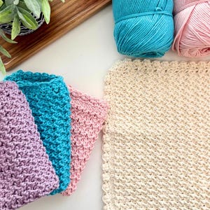 May include: A collection of handmade crochet items in various colors. The image features skeins of turquoise and pink yarn, along with crocheted washcloths in lavender, turquoise, and pink. A cream-colored square washcloth is also present. The text "I Can Crochet That" is visible.