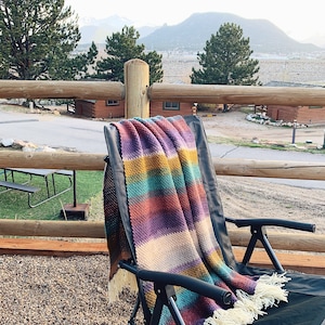 Pu&ograve; includere: Una coperta in crochet a righe colorate con frange drappeggiata su una sedia pieghevole nera. La sedia &egrave; su un patio ghiaioso con un corrimano in legno e una vista sulle montagne in lontananza.