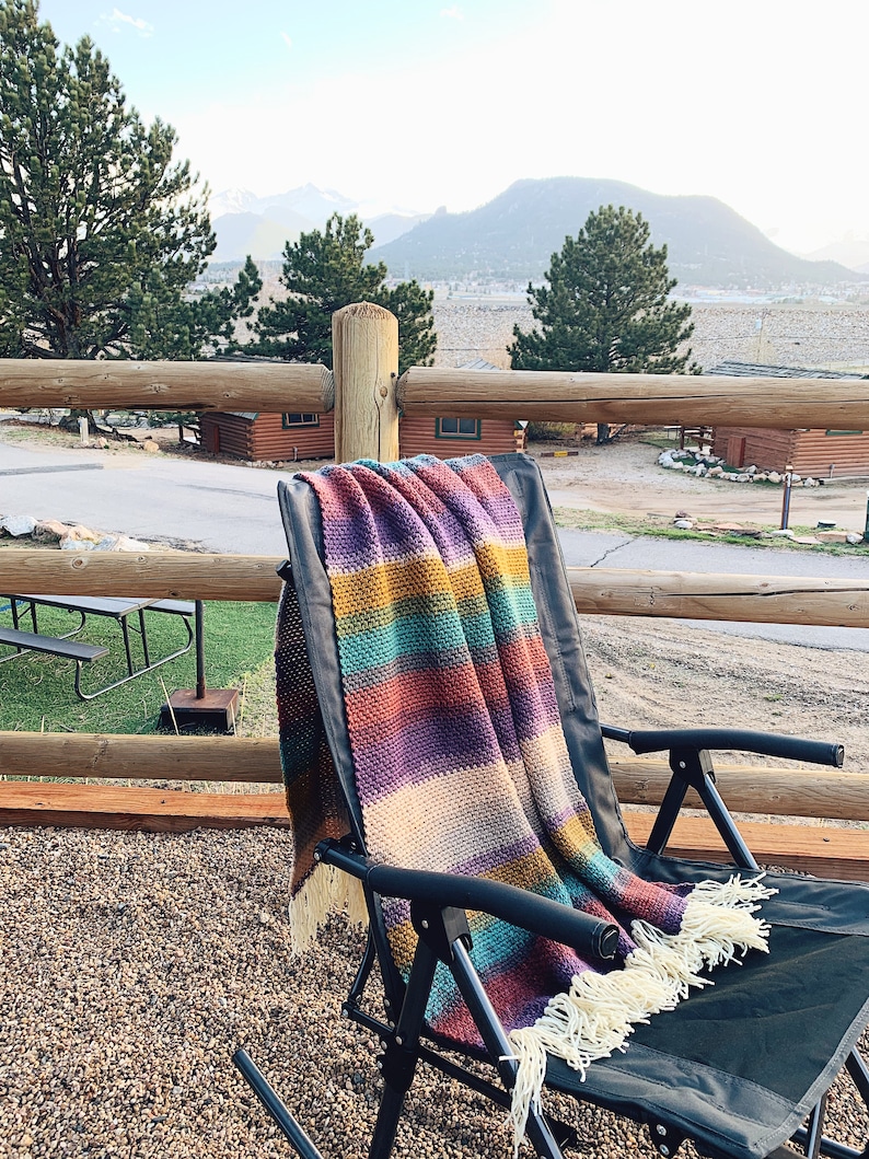 Pu&ograve; includere: Una coperta in crochet a righe colorate con frange drappeggiata su una sedia pieghevole nera. La sedia &egrave; su un patio ghiaioso con un corrimano in legno e una vista sulle montagne in lontananza.