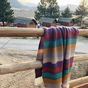 Pu&ograve; includere: Una coperta a uncinetto a righe con frange appesa a una recinzione di legno. La coperta &egrave; in tonalit&agrave; di viola, turchese, marrone e beige. Lo sfondo mostra una vista di una catena montuosa e delle capanne.