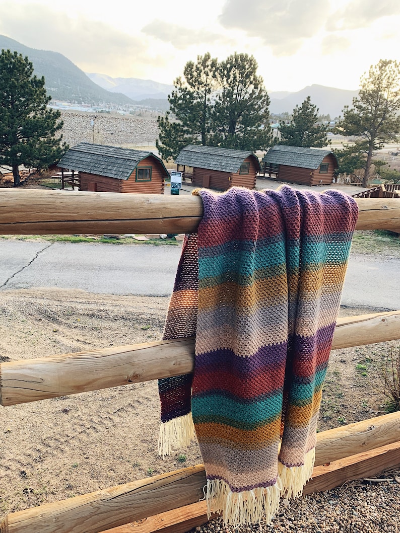 Pu&ograve; includere: Una coperta a uncinetto a righe con frange appesa a una recinzione di legno. La coperta &egrave; in tonalit&agrave; di viola, turchese, marrone e beige. Lo sfondo mostra una vista di una catena montuosa e delle capanne.