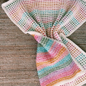 Op de afbeelding: Een kleurrijke, handgemaakte gehaakte deken. De deken heeft een wafelsteekpatroon met een regenboog aan kleuren, waaronder roze, oranje, groen en blauw. De randen zijn afgewerkt met een witte rand. De deken ligt op een getextureerd, bruin oppervlak.