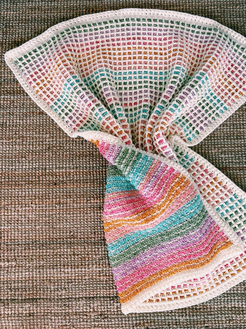 Op de afbeelding: Een kleurrijke, handgemaakte gehaakte deken. De deken heeft een wafelsteekpatroon met een regenboog aan kleuren, waaronder roze, oranje, groen en blauw. De randen zijn afgewerkt met een witte rand. De deken ligt op een getextureerd, bruin oppervlak.