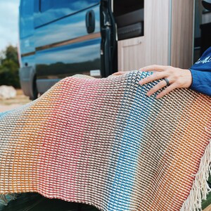 Pu&ograve; includere: Una persona &egrave; seduta su una sedia con una coperta a strisce colorate drappeggiata sulle sue ginocchia. La coperta ha un bordo a frangia ed &egrave; realizzata in un materiale morbido e tessuto. La persona indossa una camicia blu e un anello al dito. La sedia &egrave; davanti a un camper.