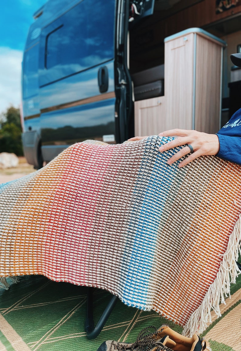 Pu&ograve; includere: Una persona &egrave; seduta su una sedia con una coperta a strisce colorate drappeggiata sulle sue ginocchia. La coperta ha un bordo a frangia ed &egrave; realizzata in un materiale morbido e tessuto. La persona indossa una camicia blu e un anello al dito. La sedia &egrave; davanti a un camper.