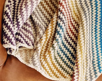 May include: A multi-colored striped crocheted throw blanket with a textured pattern. The blanket is folded and draped over a brown leather chair.