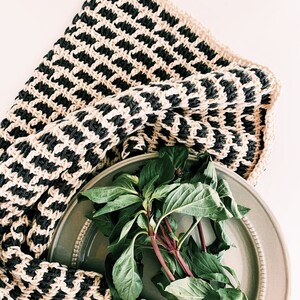 May include: A bunch of fresh green basil leaves on a gray ceramic plate with a black and cream striped crocheted dishcloth.