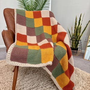 Op de afbeelding: Een handgemaakte, kleurrijke gehaakte deken over een bruine leren stoel. De deken heeft een patchwork-ontwerp met vierkanten in crème, groen, goud, oranje en bordeauxrood. De randen zijn afgewerkt met een crèmekleurige rand, wat een gezellige, getextureerde uitstraling creëert.