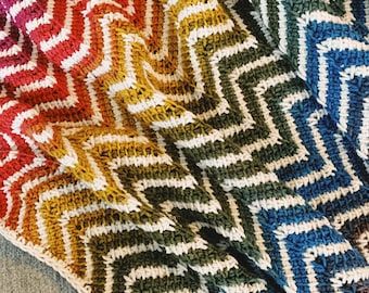 May include: A rainbow chevron crochet blanket with white stripes. The blanket is folded in half and the colors are red, orange, yellow, green, blue, and purple.