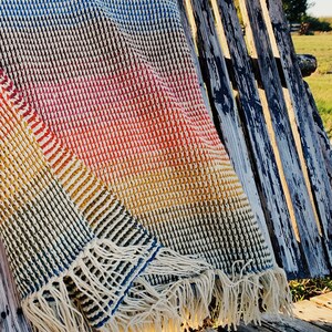 Pu&ograve; includere: Una coperta in crochet multicolore con un bordo a frangia. La coperta &egrave; drappeggiata su una panca di legno invecchiato.