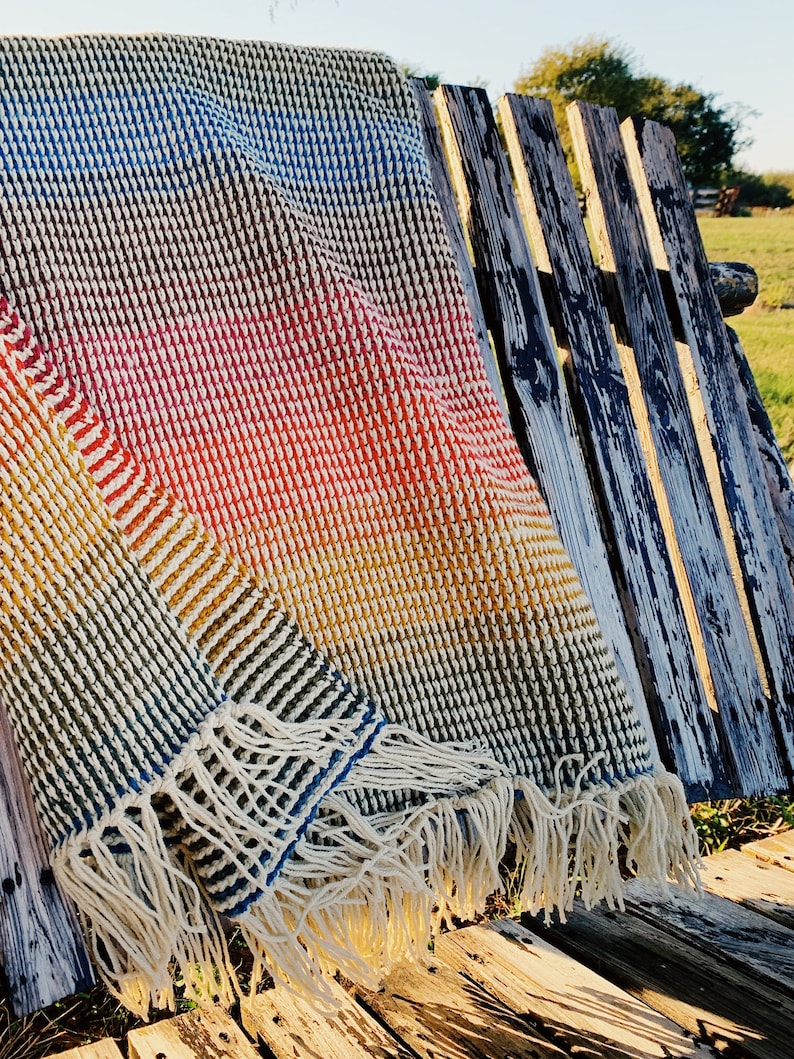 Pu&ograve; includere: Una coperta in crochet multicolore con un bordo a frangia. La coperta &egrave; drappeggiata su una panca di legno invecchiato.