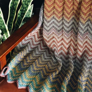Pu&ograve; includere: Una coperta all'uncinetto con motivo chevron multicolore in tonalit&agrave; di marrone, rosso, viola, blu, verde e giallo. La coperta &egrave; drappeggiata su una sedia di legno.