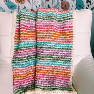 Op de afbeelding: Een kleurrijke, gestreepte gehaakte deken in tinten roze, oranje, groen en blauw. De deken heeft een cr&egrave;mekleurige rand en is over een witte stoel gedrapeerd. Het ontwerp van de deken heeft horizontale strepen.