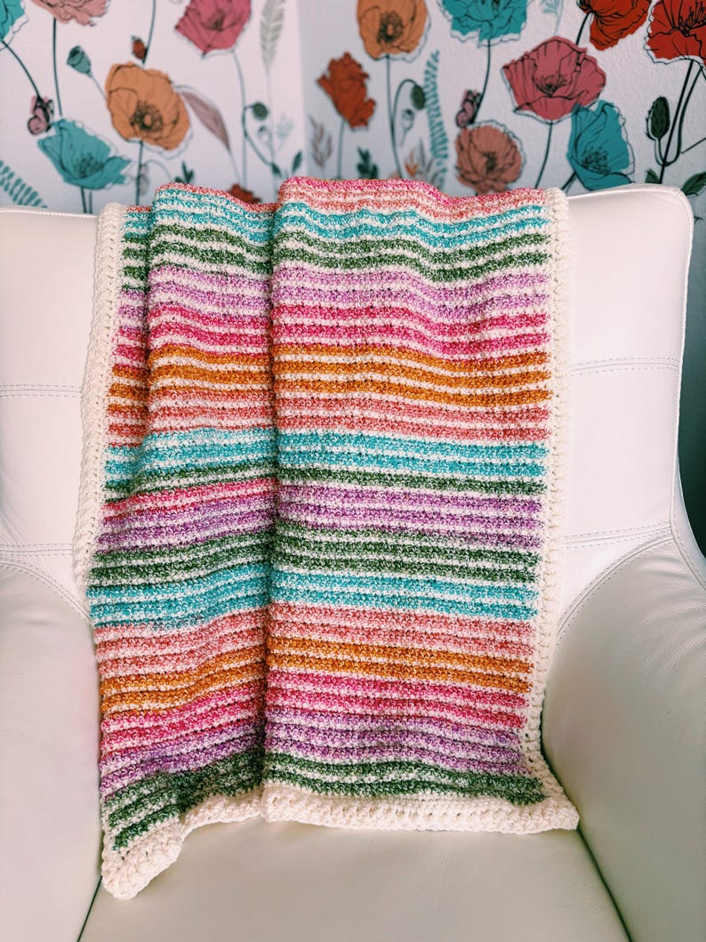 Op de afbeelding: Een kleurrijke, gestreepte gehaakte deken in tinten roze, oranje, groen en blauw. De deken heeft een cr&egrave;mekleurige rand en is over een witte stoel gedrapeerd. Het ontwerp van de deken heeft horizontale strepen.