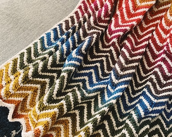 May include: A colorful chevron crochet blanket with a white border. The blanket is made with a variety of colors, including red, orange, yellow, green, blue, and purple.
