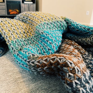 Puede incluir: Una manta de crochet multicolor con rayas de color amarillo, verde, azul y marr&oacute;n. La manta est&aacute; colocada sobre un sof&aacute;.