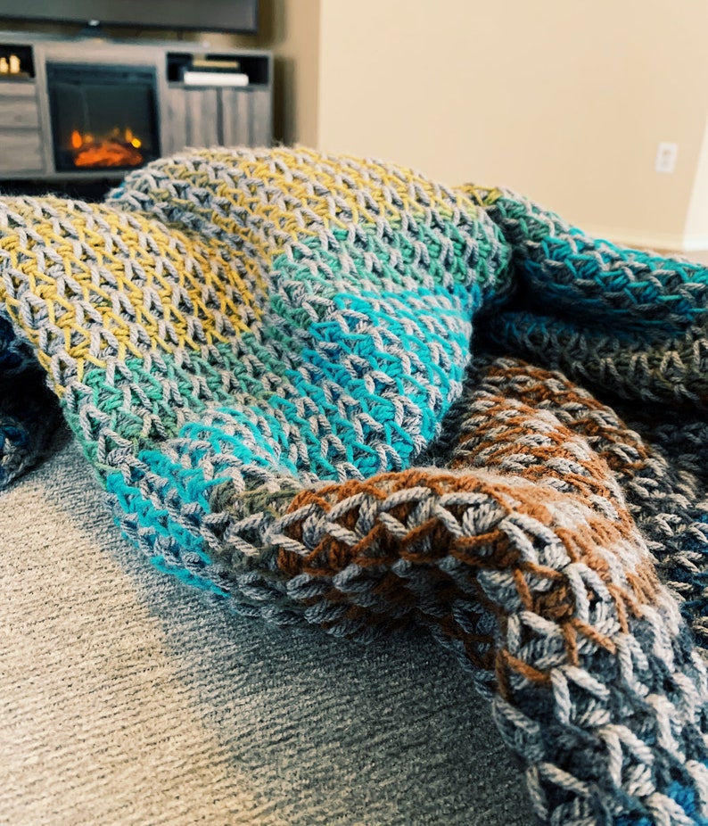 Puede incluir: Una manta de crochet multicolor con rayas de color amarillo, verde, azul y marr&oacute;n. La manta est&aacute; colocada sobre un sof&aacute;.