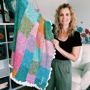 May include: A vibrant, handmade blanket with a patchwork design. The blanket features squares of pink, orange, green, blue, and purple. It has a white, scalloped edge and a monogrammed initial. The blanket is made of a soft, textured material.