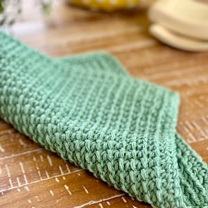 K&ouml;nnte beinhalten: Ein weiches, gr&uuml;nes, gestricktes Geschirrtuch oder Teetuch. Das Tuch ist in der H&auml;lfte gefaltet und liegt auf einer Holzoberfl&auml;che.