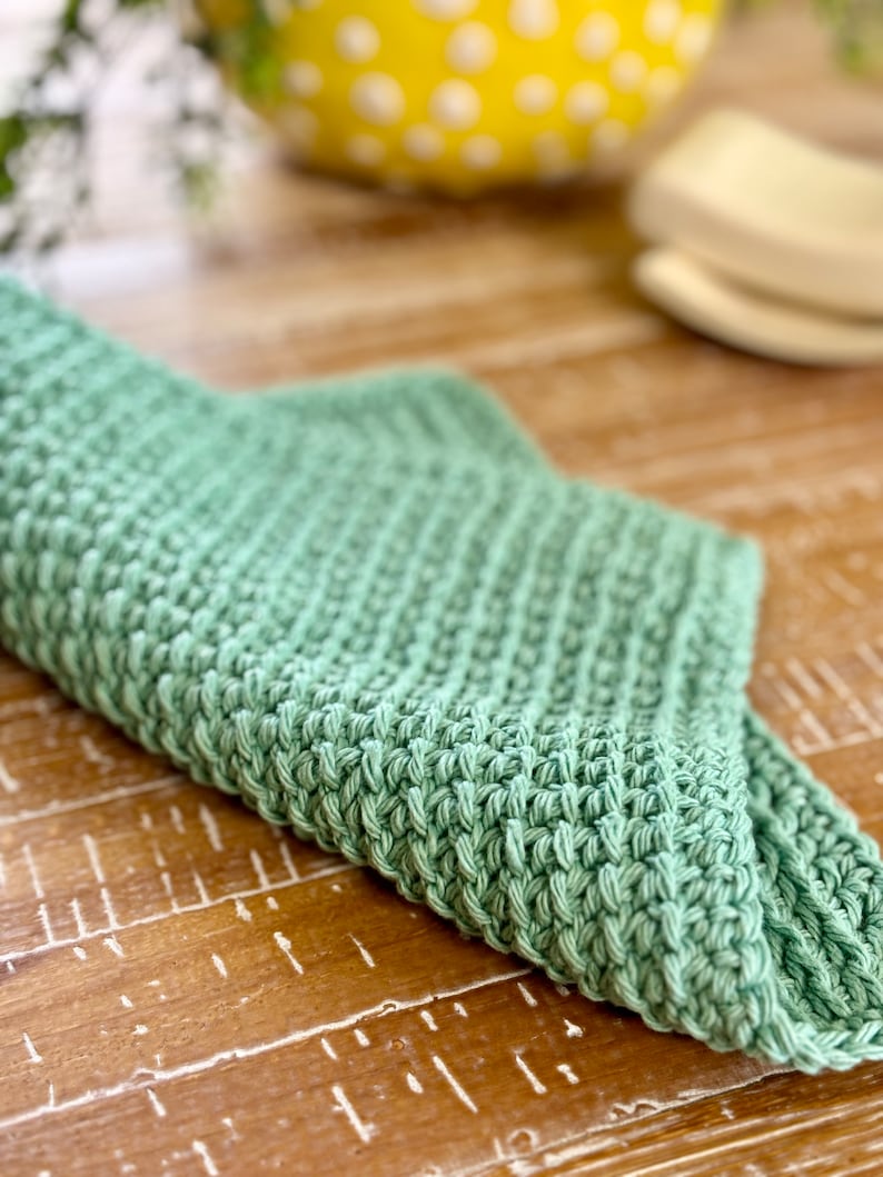 K&ouml;nnte beinhalten: Ein weiches, gr&uuml;nes, gestricktes Geschirrtuch oder Teetuch. Das Tuch ist in der H&auml;lfte gefaltet und liegt auf einer Holzoberfl&auml;che.
