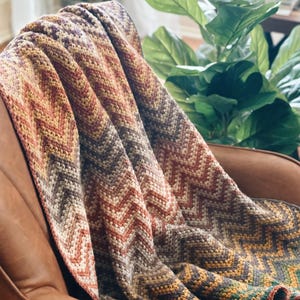 Pu&ograve; includere: Una coperta all'uncinetto con un motivo a chevron in tonalit&agrave; di marrone, arancio, giallo e verde. La coperta &egrave; drappeggiata su una poltrona in pelle marrone.