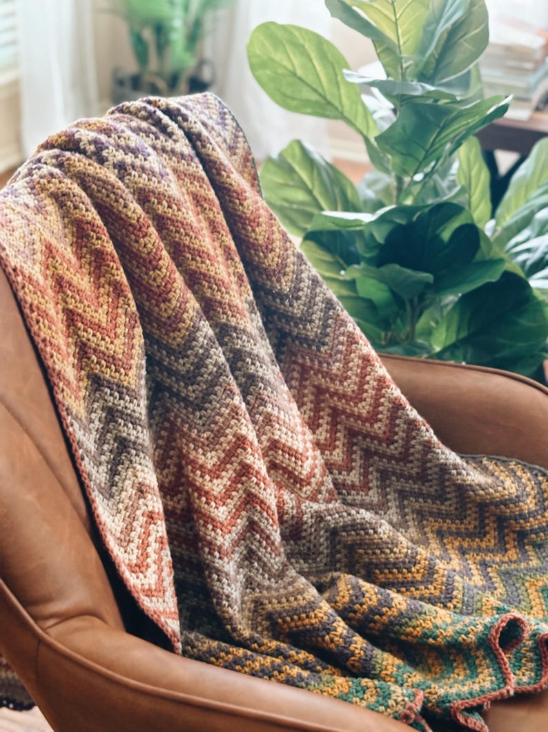 Pu&ograve; includere: Una coperta all'uncinetto con un motivo a chevron in tonalit&agrave; di marrone, arancio, giallo e verde. La coperta &egrave; drappeggiata su una poltrona in pelle marrone.