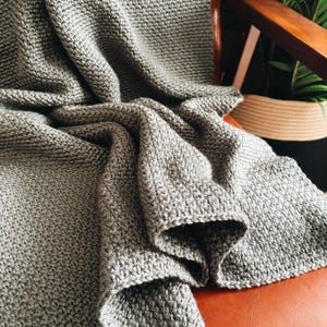 Op de afbeelding: Een grijze gebreide deken gedrapeerd over een bruine leren stoel. De deken is zacht en heeft een textuur, met een subtiele glans.