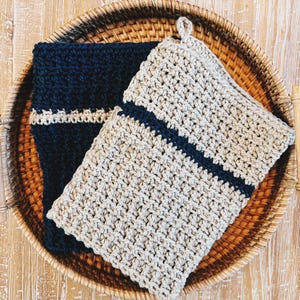 Puede incluir: Dos agarraderas de olla de ganchillo en una cesta tejida. Una es beige con una franja azul oscuro y la otra es azul oscuro con una franja beige.