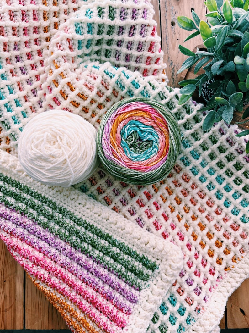 Op de afbeelding: Een cr&egrave;mekleurige gehaakte deken met wafelsteek met kleurrijke accenten van roze, oranje, groen en blauw. Twee bollen garen liggen naast de deken, &eacute;&eacute;n wit en &eacute;&eacute;n veelkleurig. De deken is op een houten oppervlak gevouwen.