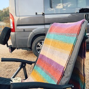 Pu&ograve; includere: Una coperta all'uncinetto a righe drappeggiata su una sedia pieghevole nera davanti a un furgone grigio.