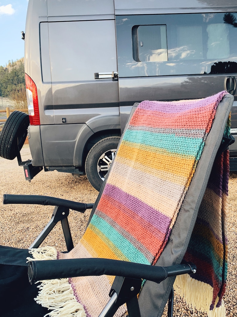 Pu&ograve; includere: Una coperta all'uncinetto a righe drappeggiata su una sedia pieghevole nera davanti a un furgone grigio.