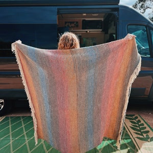 Pu&ograve; includere: Una persona tiene in mano una coperta di lana tessuta a righe colorate con frange. La coperta &egrave; davanti a un furgone nero con la porta aperta.