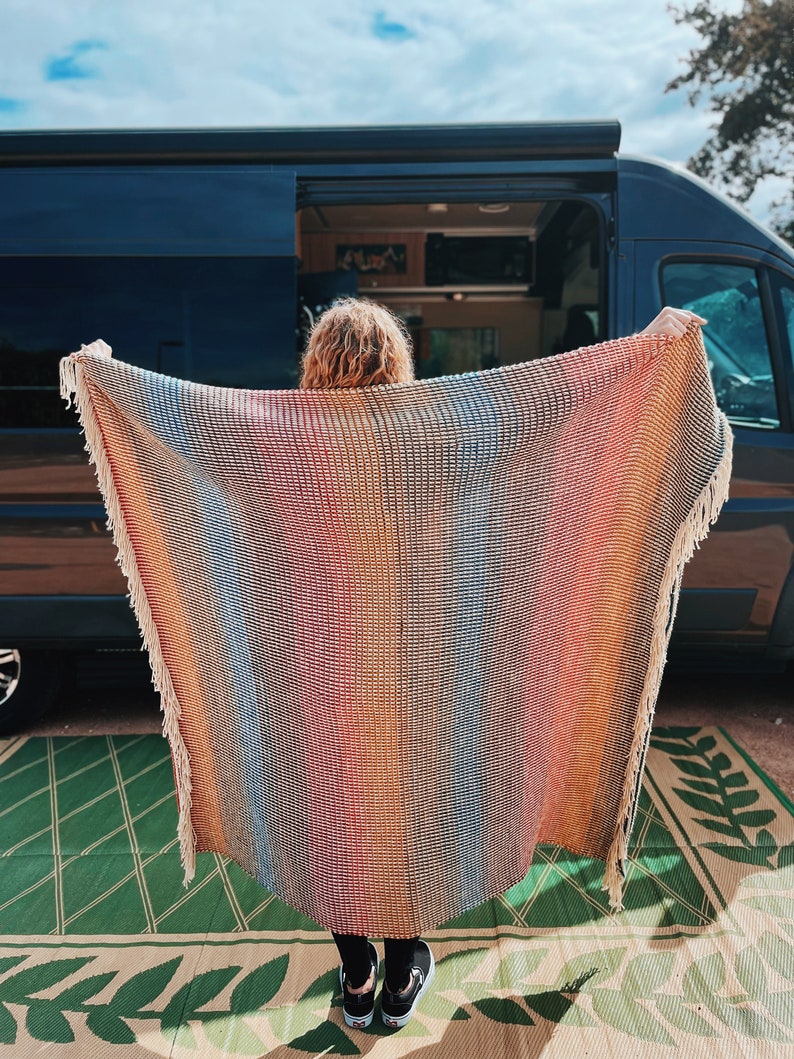 Pu&ograve; includere: Una persona tiene in mano una coperta di lana tessuta a righe colorate con frange. La coperta &egrave; davanti a un furgone nero con la porta aperta.