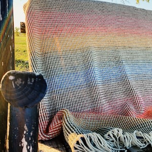 Pu&ograve; includere: Una coperta in crochet a righe arcobaleno con frange su una panca di legno.