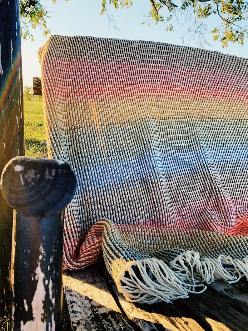 Pu&ograve; includere: Una coperta in crochet a righe arcobaleno con frange su una panca di legno.