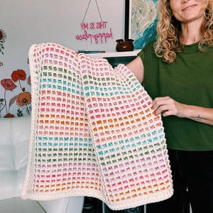 Op de afbeelding: Een handgemaakte, kleurrijke gehaakte deken met een wafelsteekpatroon. De deken heeft een kleurengradient, waaronder roze, oranje, groen en blauw, met een cr&egrave;mekleurige rand. De deken wordt voor een witte stoel gehouden.