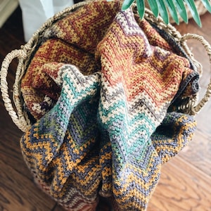 Pu&ograve; includere: Un cesto intrecciato pieno di una coperta all'uncinetto colorata con un motivo a chevron in tonalit&agrave; di marrone, arancione, verde acqua e grigio.