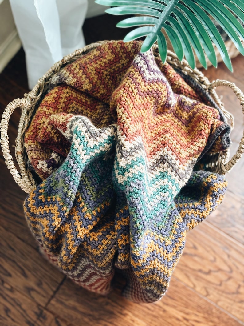Pu&ograve; includere: Un cesto intrecciato pieno di una coperta all'uncinetto colorata con un motivo a chevron in tonalit&agrave; di marrone, arancione, verde acqua e grigio.