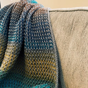 Puede incluir: Una manta de ganchillo multicolor con tonos de azul, amarillo y marr&oacute;n. La manta est&aacute; drapeada sobre un sof&aacute; gris.