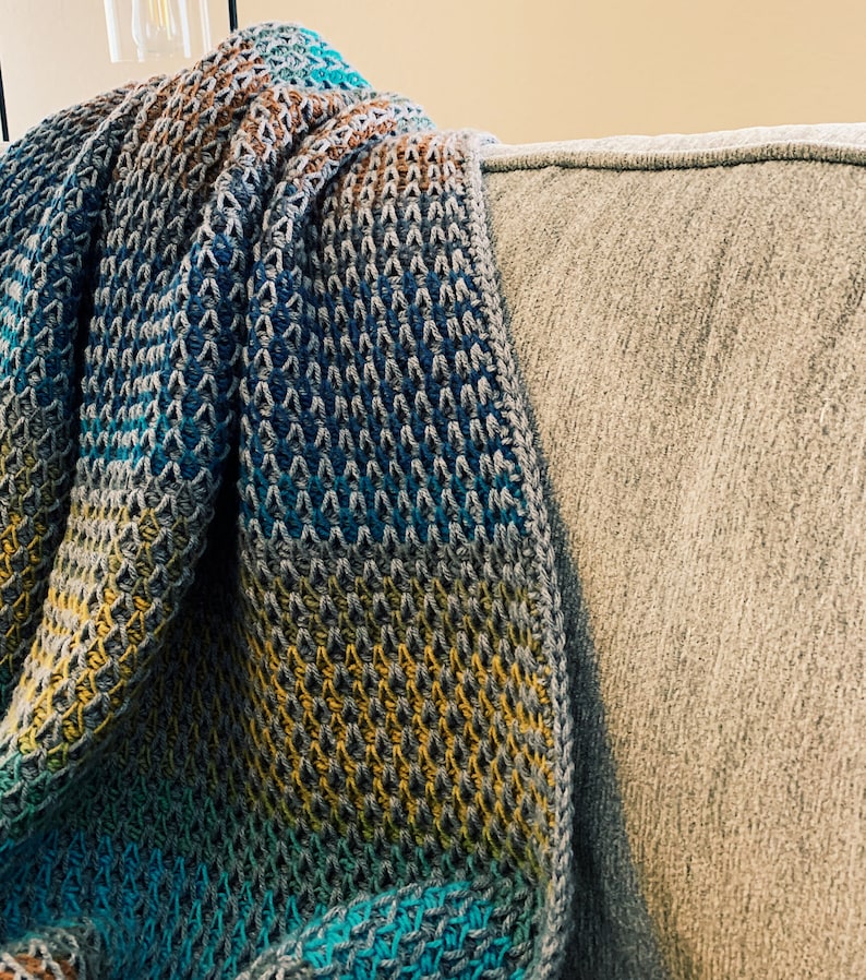 Puede incluir: Una manta de ganchillo multicolor con tonos de azul, amarillo y marr&oacute;n. La manta est&aacute; drapeada sobre un sof&aacute; gris.