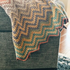 Pu&ograve; includere: Una coperta all'uncinetto con un motivo a chevron in tonalit&agrave; di marrone, arancione, verde e viola. La coperta &egrave; piegata e drappeggiata su un divano.