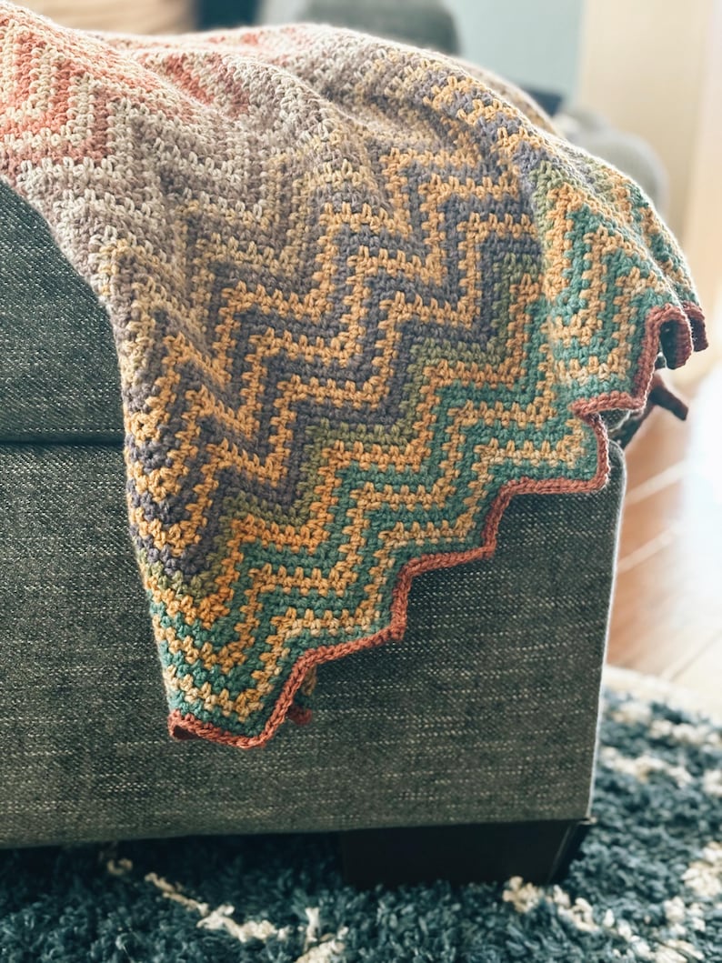 Pu&ograve; includere: Una coperta all'uncinetto con un motivo a chevron in tonalit&agrave; di marrone, arancione, verde e viola. La coperta &egrave; piegata e drappeggiata su un divano.